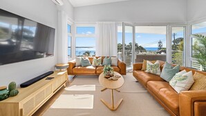A spacious living room with a large flat-screen TV, two leather sofas, a coffee table, and a large window offering a view of the ocean. - Tea Trees Ocean Views, Pet-Friendly, Walk to shops and Beach (Woolgoolga)