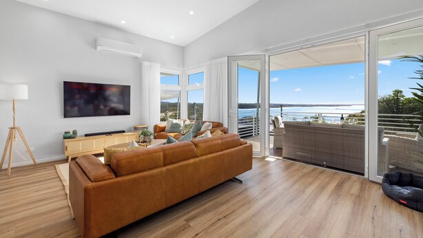 A spacious living room with a large sofa, a flat-screen TV, and a view of the ocean through large sliding doors. - Tea Trees Ocean Views, Pet-Friendly, Walk to shops and Beach (Woolgoolga)