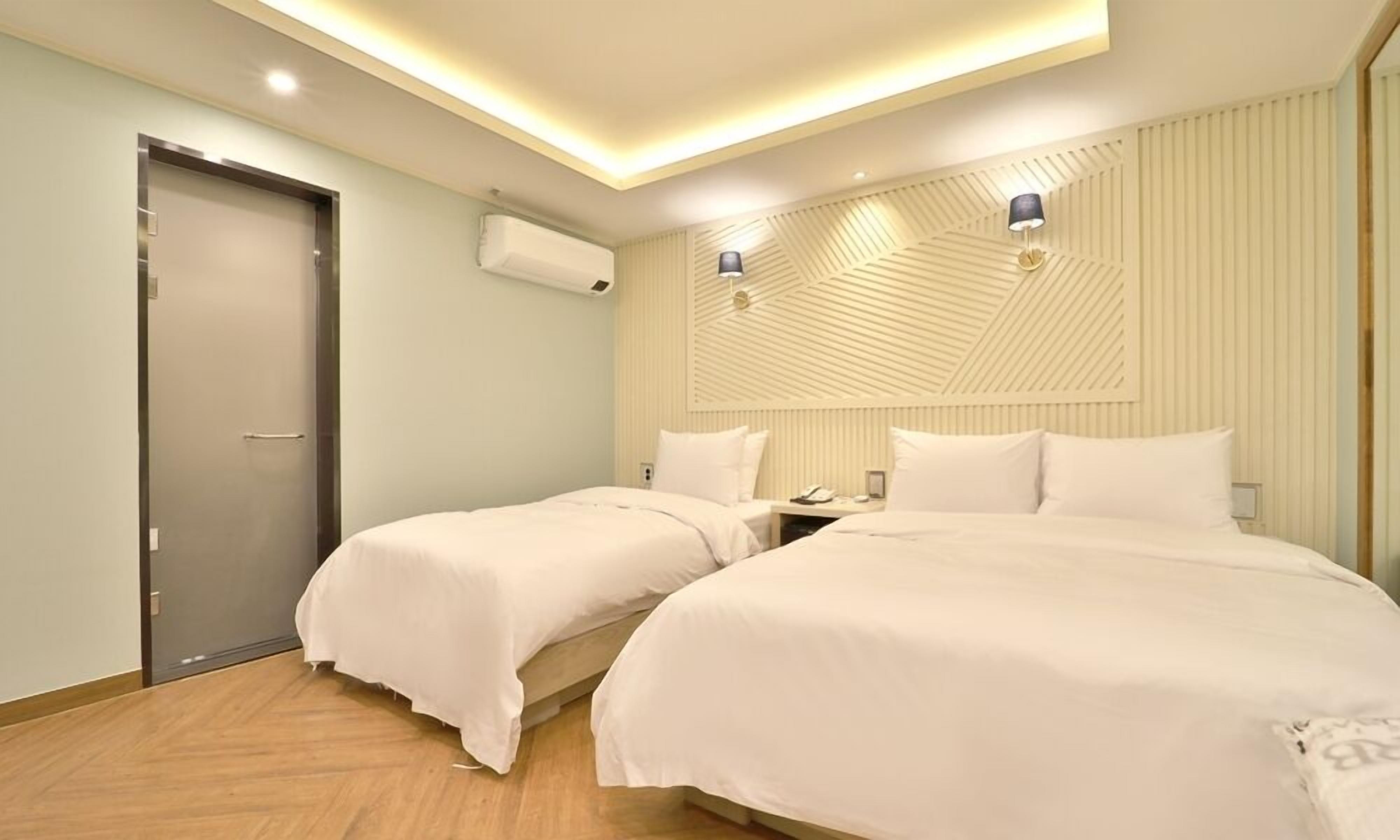 A hotel room with two beds, a door, an air conditioning unit, and a wall-mounted light fixture.