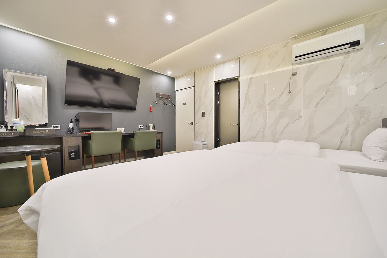 A modern hotel room with a large bed, a desk with a chair, a flat-screen TV, a mirror, and an air conditioning unit.