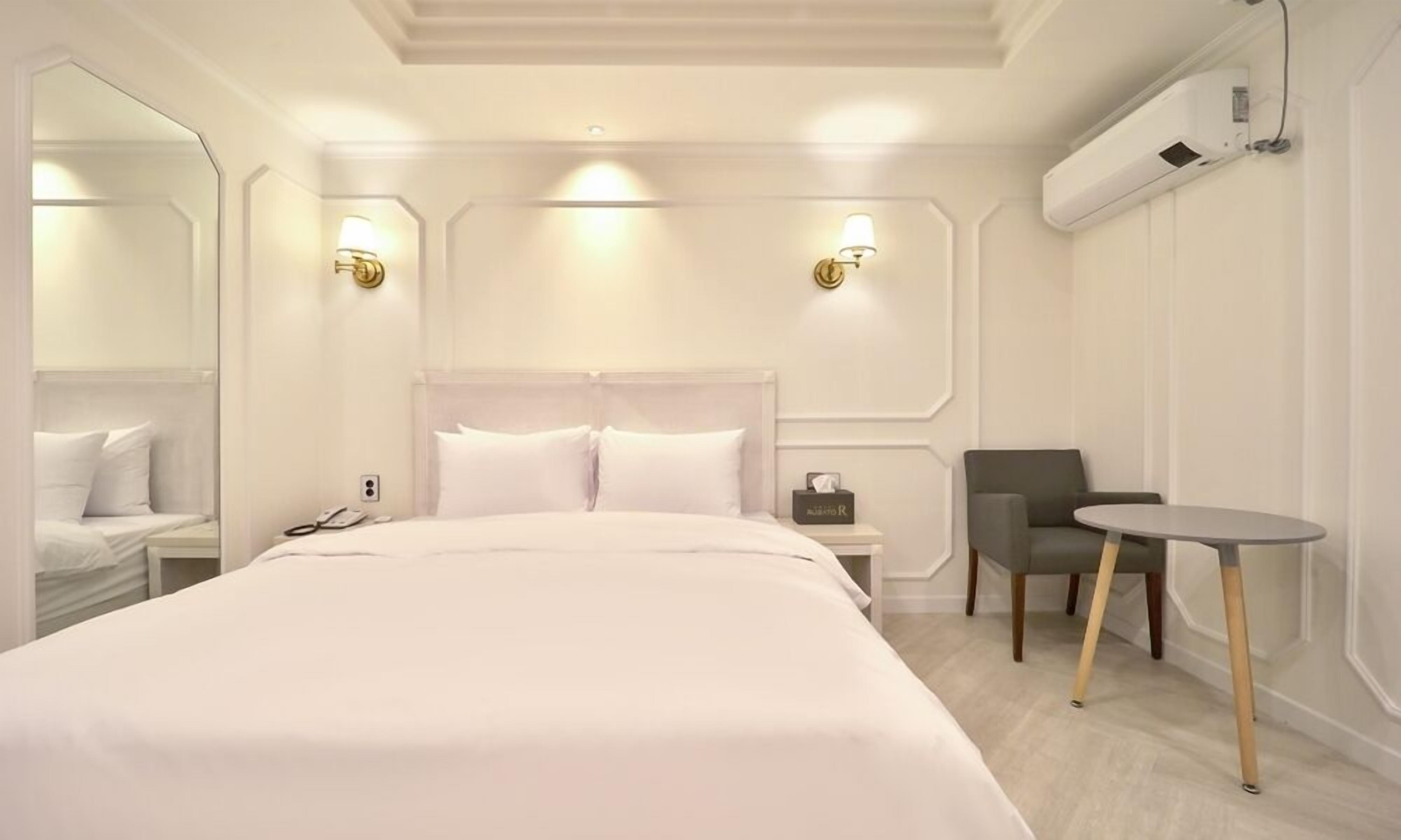 A hotel room with a large bed, bedside tables, a chair, a small table, and a mirror.