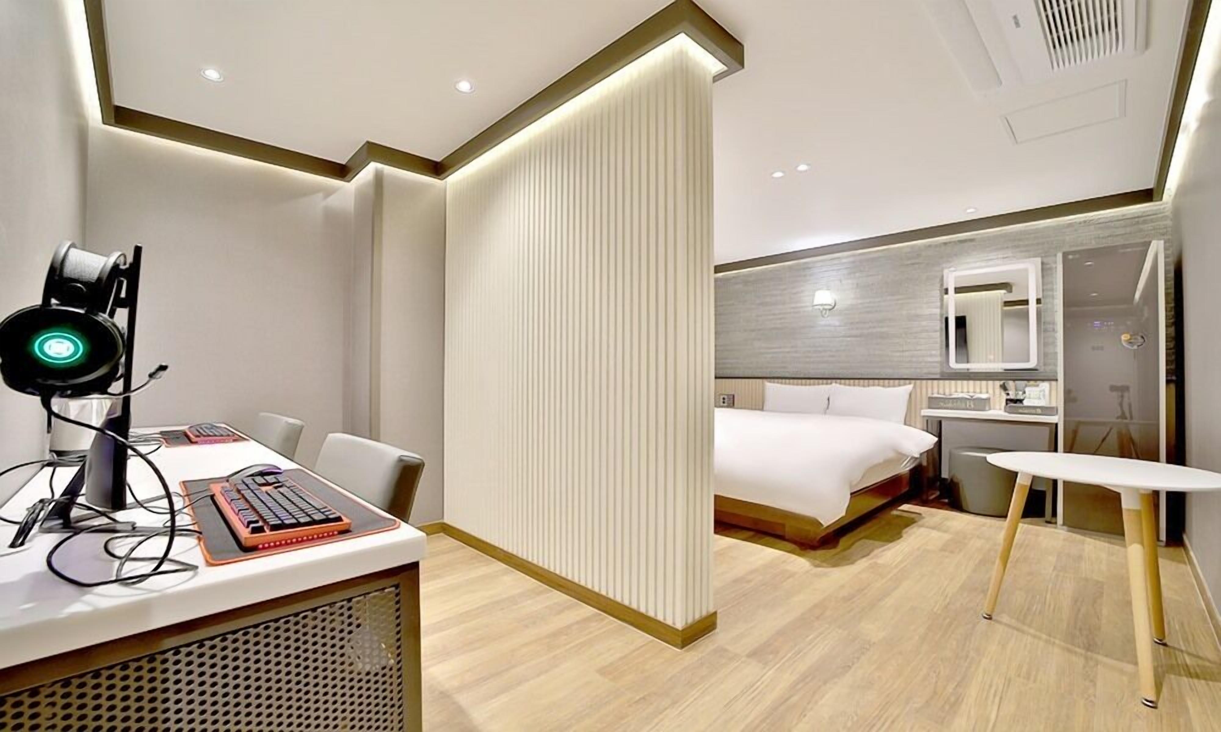 A modern hotel room with a desk, computer, headphones, and a bed.