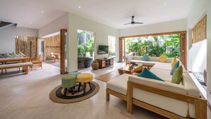 A spacious living room with a sofa, ottoman, and dining area. There is a large window with a view of greenery outside. - Puri Aman - Unique & Lush 3BR Pool Villa - Next to Gigi Susu & Walk to Beach (Canggu)