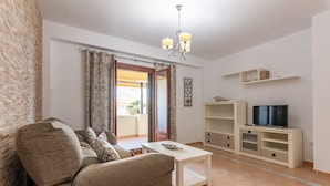 Una sala de estar con un sofá, una mesa de centro, un televisor sobre un mueble bajo y un balcón con cortinas.