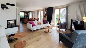 Een ruime woonkamer met een witte bank, een zwarte fauteuil en een televisie. Er is een eethoek met een tafel en stoelen, en een keuken is zichtbaar op de achtergrond.