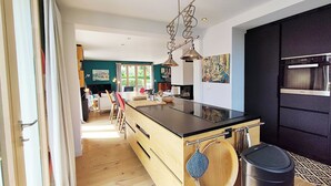 Een moderne keuken met een centraal eiland, houten kastjes en een zwart aanrecht. Er hangt een zwarte prullenbak en een blauwe waterkoker aan het eiland.