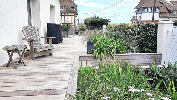 Een houten terras met tuinmeubilair, een tuin met verschillende planten, en een wit gebouw met raam en deur.
