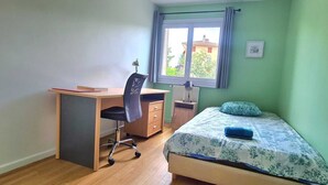 Un dormitorio con cama, escritorio y silla. Se ve una ventana con vistas a edificios.