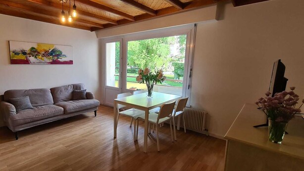 Una sala de estar acogedora con techo de madera, un sofá gris, una mesa de comedor con sillas y una ventana con vistas a zonas verdes.
