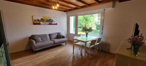 Una sala de estar acogedora con techo de madera, un sofá gris, una mesa de comedor con sillas y una ventana con vistas a zonas verdes.