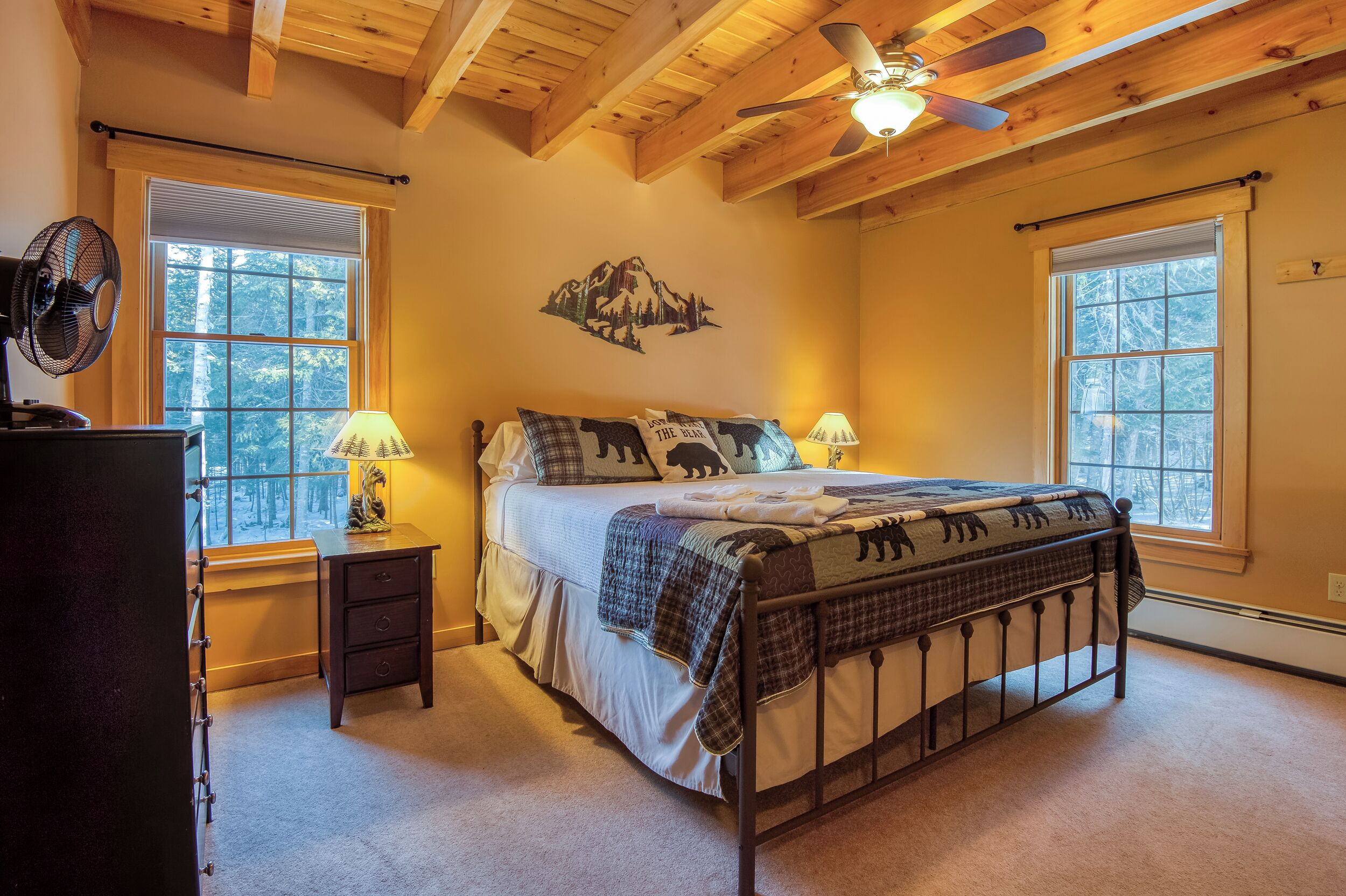 A bedroom with a bed, two bedside lamps, a wall-mounted fan, and a window with a view of trees.