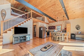 A spacious living area with a wooden staircase, a large flat-screen TV, a dining table, and a kitchen in the background. - Bear Pause - 2 bedroom, 2 bath close to downtown Rangeley! (Rangeley)