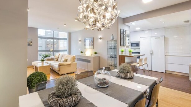 Ein modernes Wohnzimmer mit Essbereich, ausgestattet mit einer Chandelier, Sofas und einer Küche im Hintergrund.