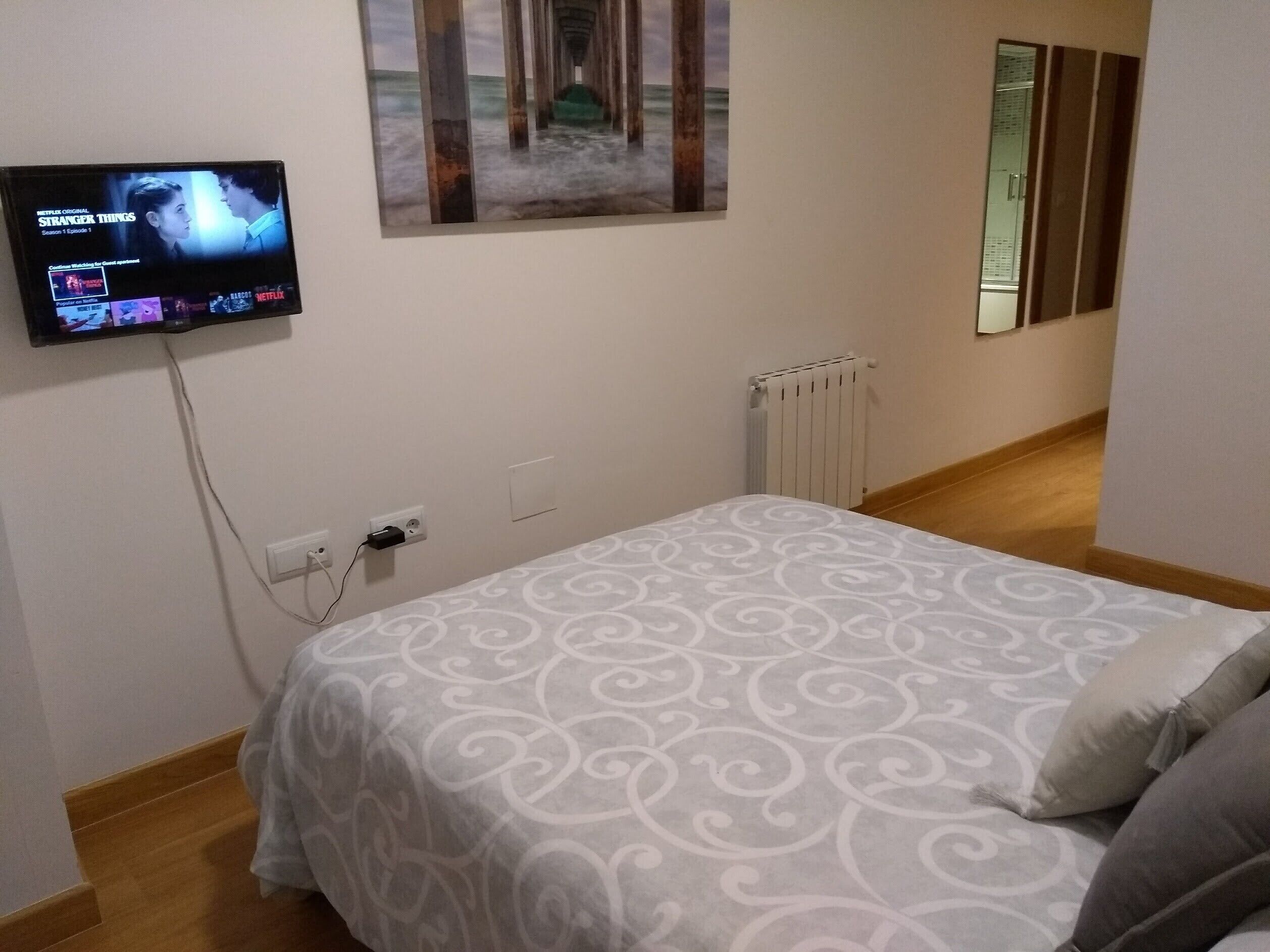 A bedroom with a bed, a TV mounted on the wall, a painting, and a radiator.