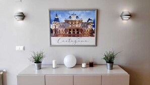 Ein gerahmtes Kunstwerk mit einem Gebäude und dem Schriftzug „Cartagena“ darunter, platziert über einem weißen Konsolentisch mit zwei Topfpflanzen, zwei Kerzen und einer kugelförmigen Vase.