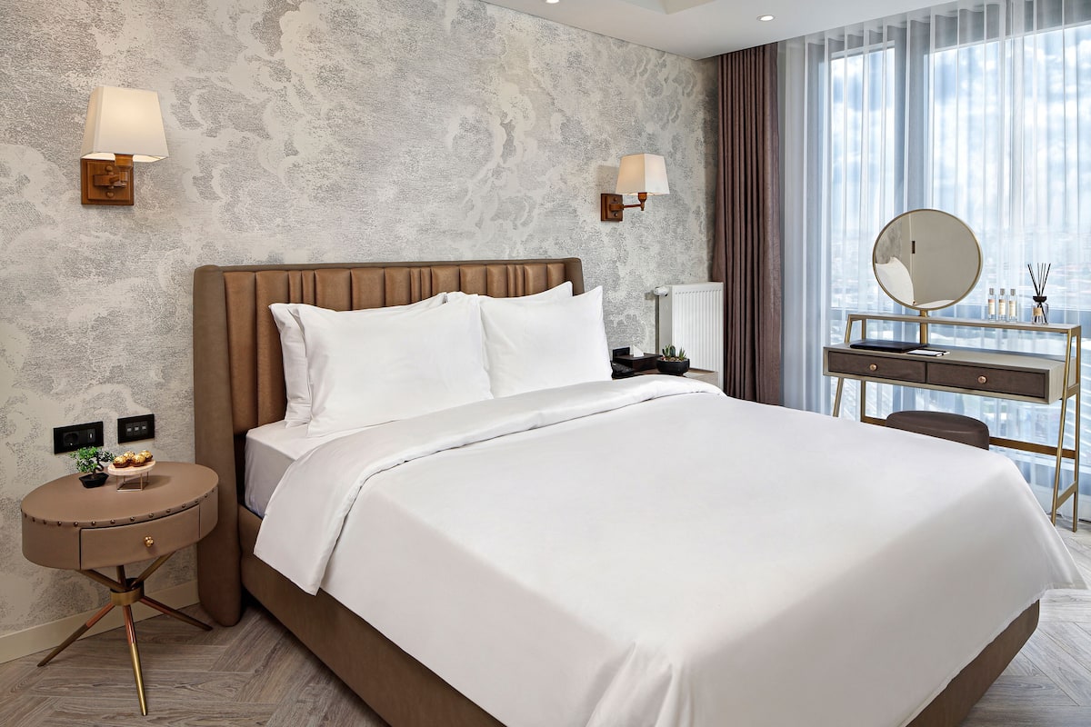 A modern hotel room with a large bed, two bedside lamps, a round mirror, a small table with a plant, and a view of the city through large windows.