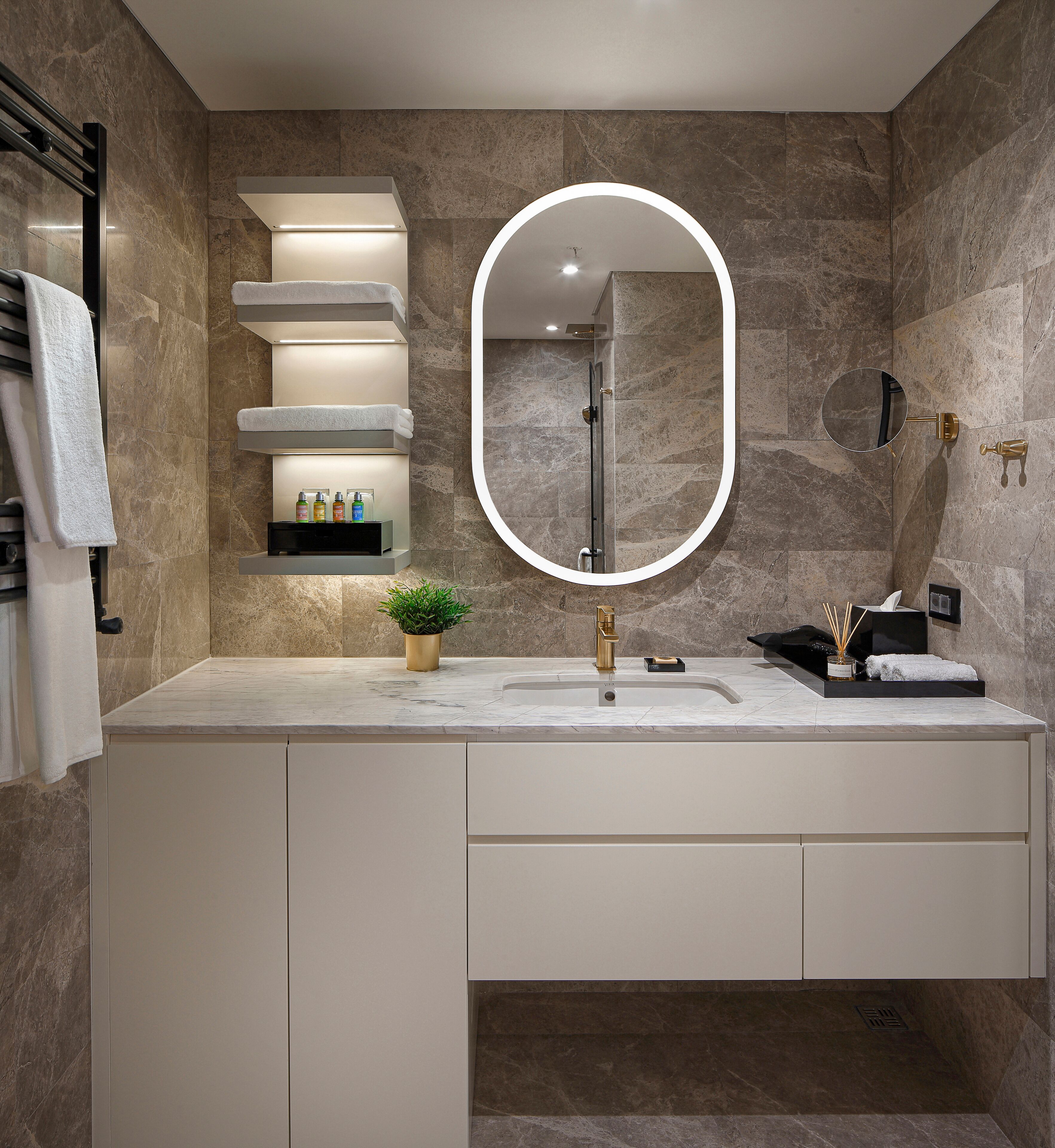 Un baño moderno con pared de mármol, un espejo ovalado grande, un grifo dorado y un estante empotrado con artículos de aseo.