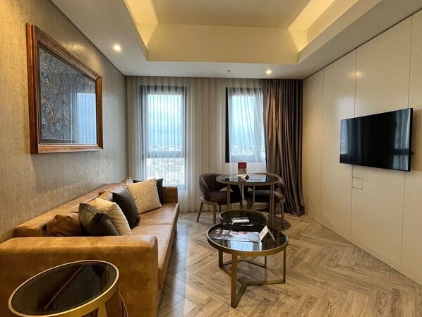 A modern hotel room with a leather sofa, a round dining table, and a flat-screen TV mounted on the wall.