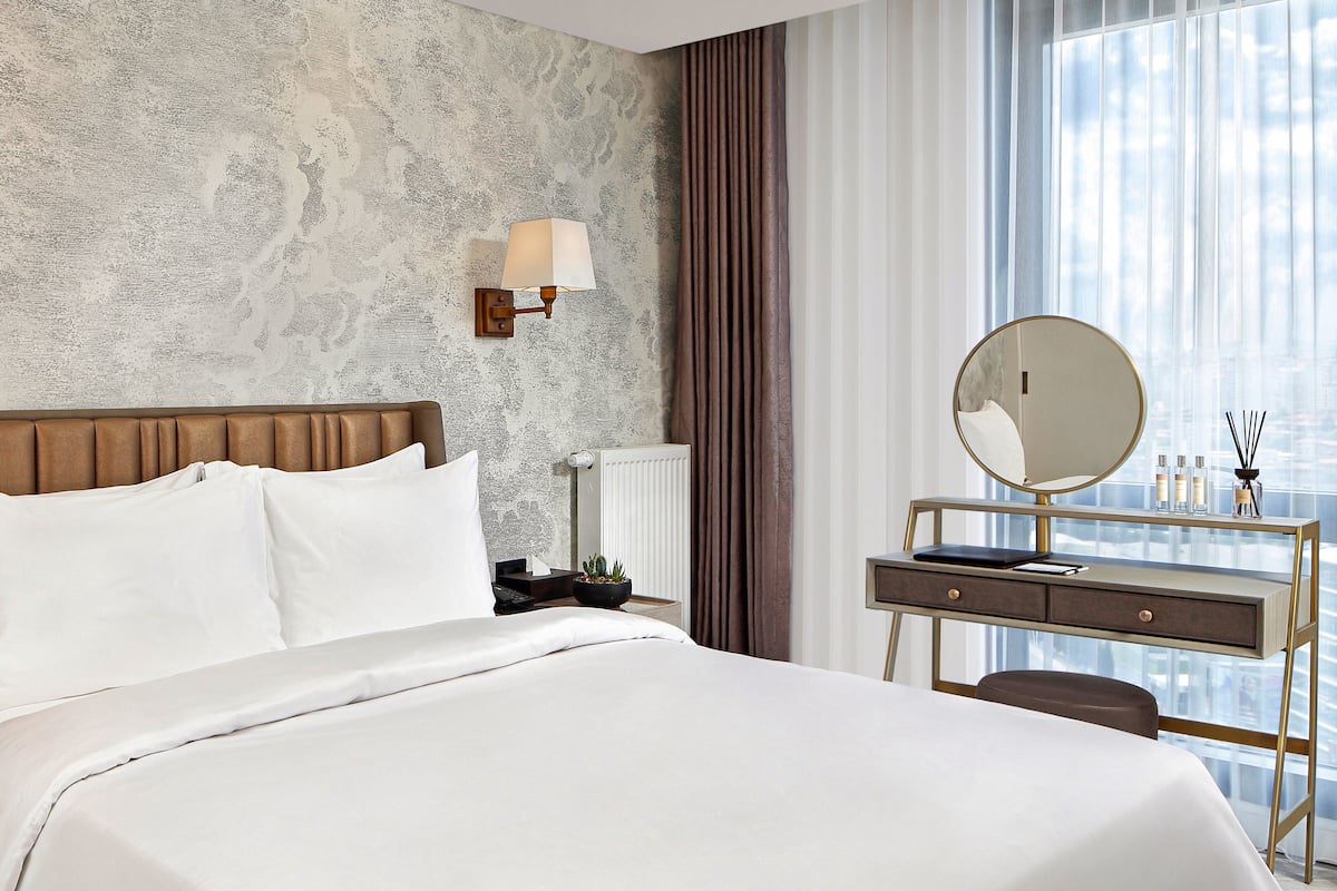 A hotel room with a large bed, a bedside table with a lamp, a dresser with a mirror, and a window with curtains.