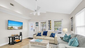 A modern living room with a beige sofa, a grey armchair, and a coffee table. There is a television mounted on the wall, a ceiling fan, and a window with blinds. - West Destin!  3 blks to beach! Front & back porch! Walk to shopping/restaurants (Destin)