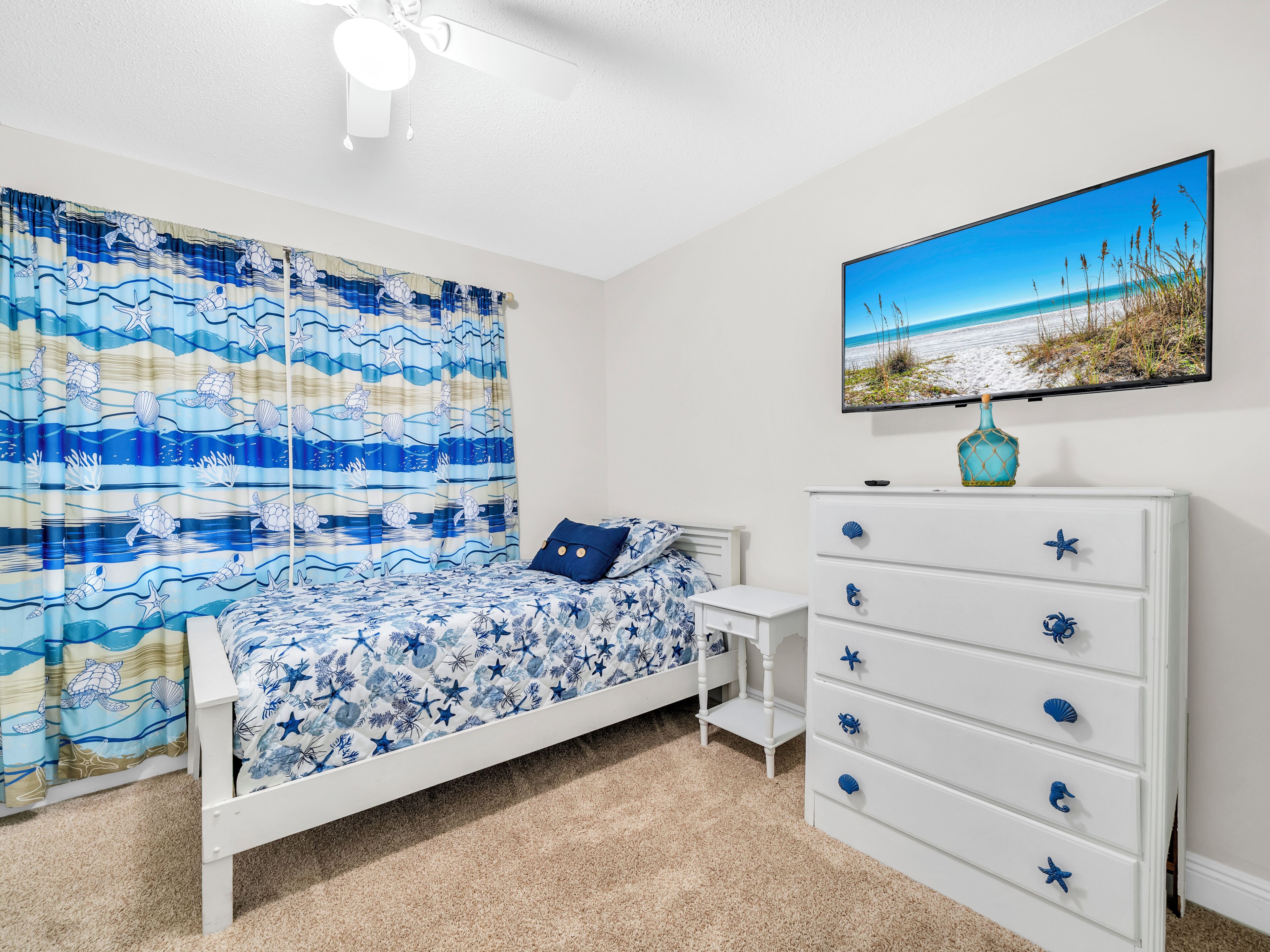 Ein Schlafzimmer mit Bett, Kommode, Nachttisch, Deckenventilator und einem Strandbild.