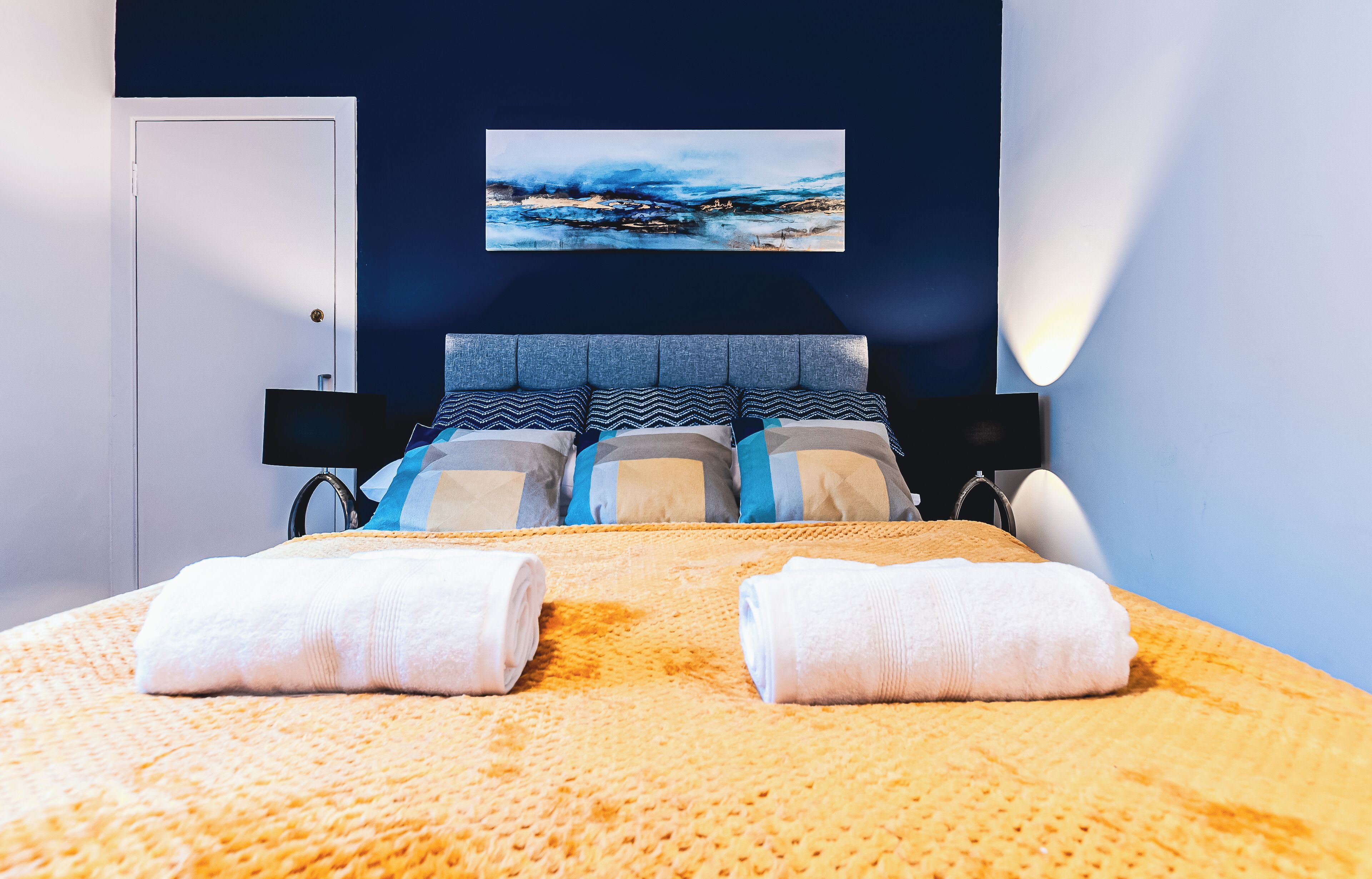 A neatly made bed with two towels, a headboard with a painting, and a bedside lamp.