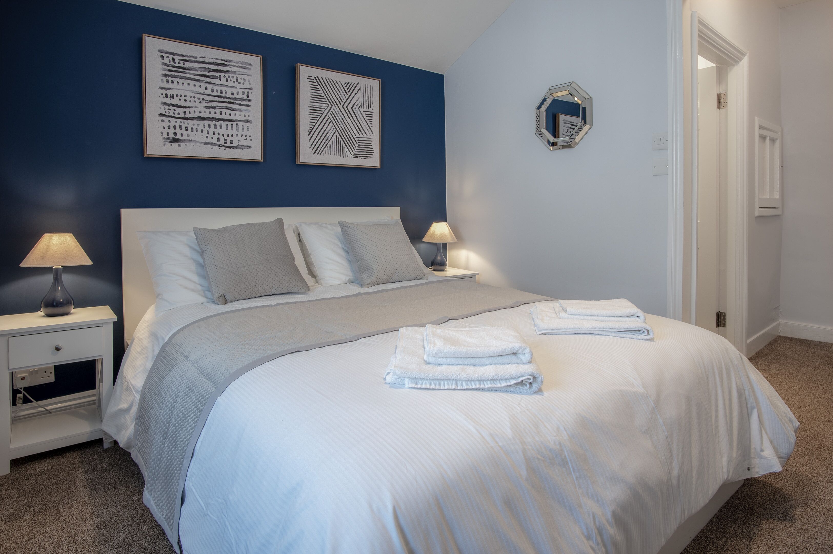 A neatly made bed with white linens and folded towels, a bedside table with a lamp, and two wall art pieces.