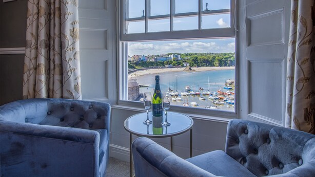 A room with two blue armchairs and a small round table with a bottle and glasses. A large window offers a view of a beach and boats. - Gwynne House - 6 Bedroom - Tenby Harbour (Tenby)