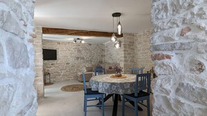 Un comedor con pared de piedra, una mesa redonda con mantel floral, sillas azules y lámparas colgantes.