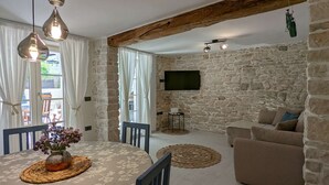 Una sala de estar con pared de piedra, un televisor montado en la pared, un sofá, una mesa de centro con un jarrón de flores y un comedor con una mesa y sillas.