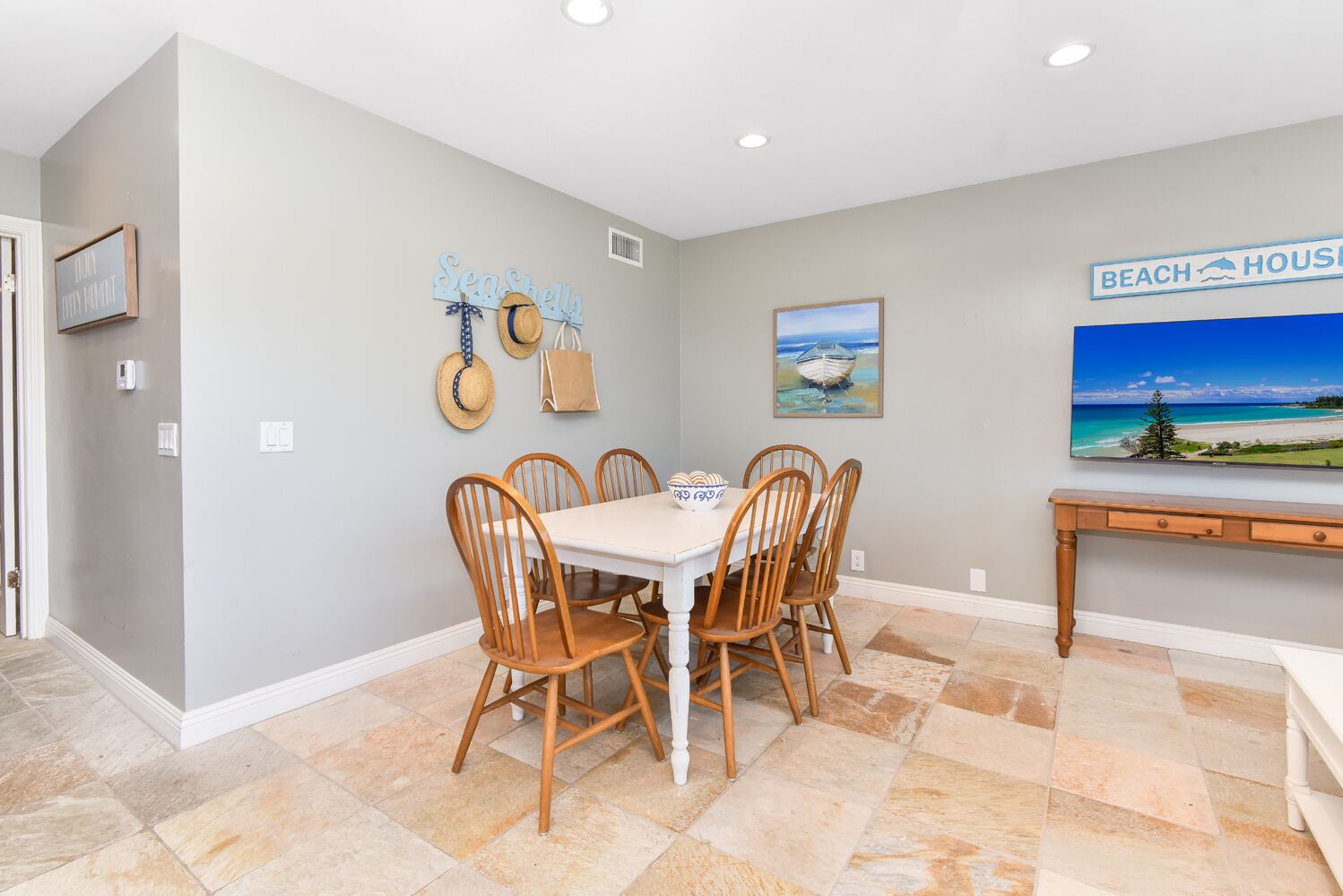 Un comedor con una mesa blanca y sillas de madera, un cuadro de una playa y un letrero en la pared que dice 'BEACH HOUSE'.