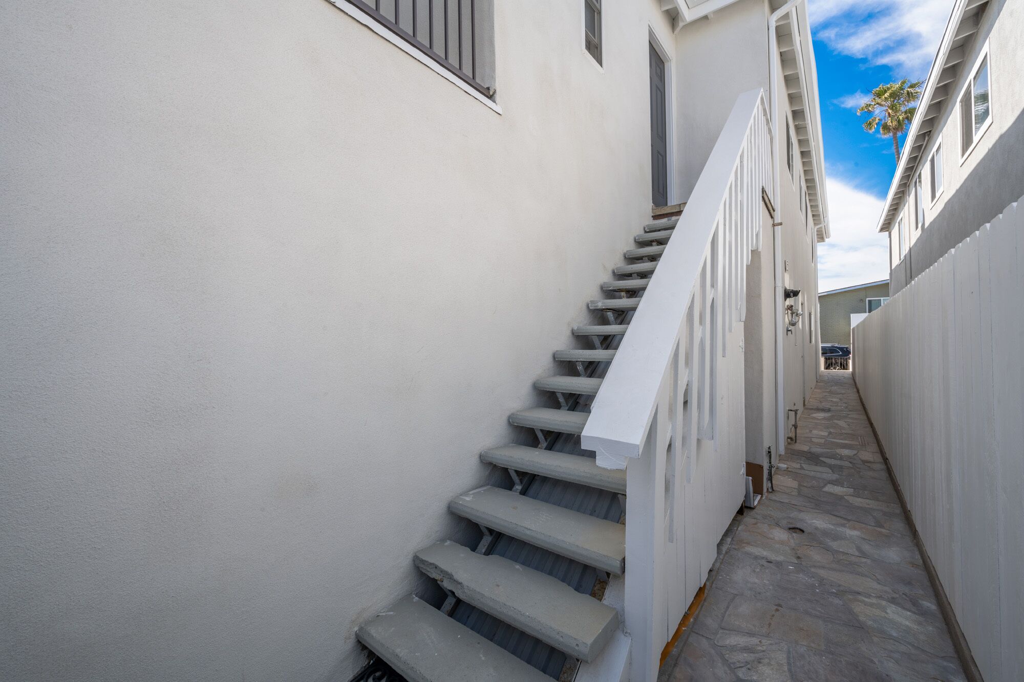 Un escalier étroit aux murs blancs, doté d’une rampe métallique, mène à une porte située en haut.