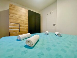 Habitación de hotel con cama, dos toallas enrolladas, armario de madera y puerta blanca con pomo.