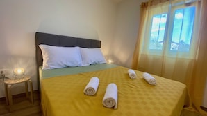 Un lit bien fait, avec une couvre-lit jaune et des oreillers blancs, une petite table de chevet avec une lampe, et une fenêtre avec des rideaux.