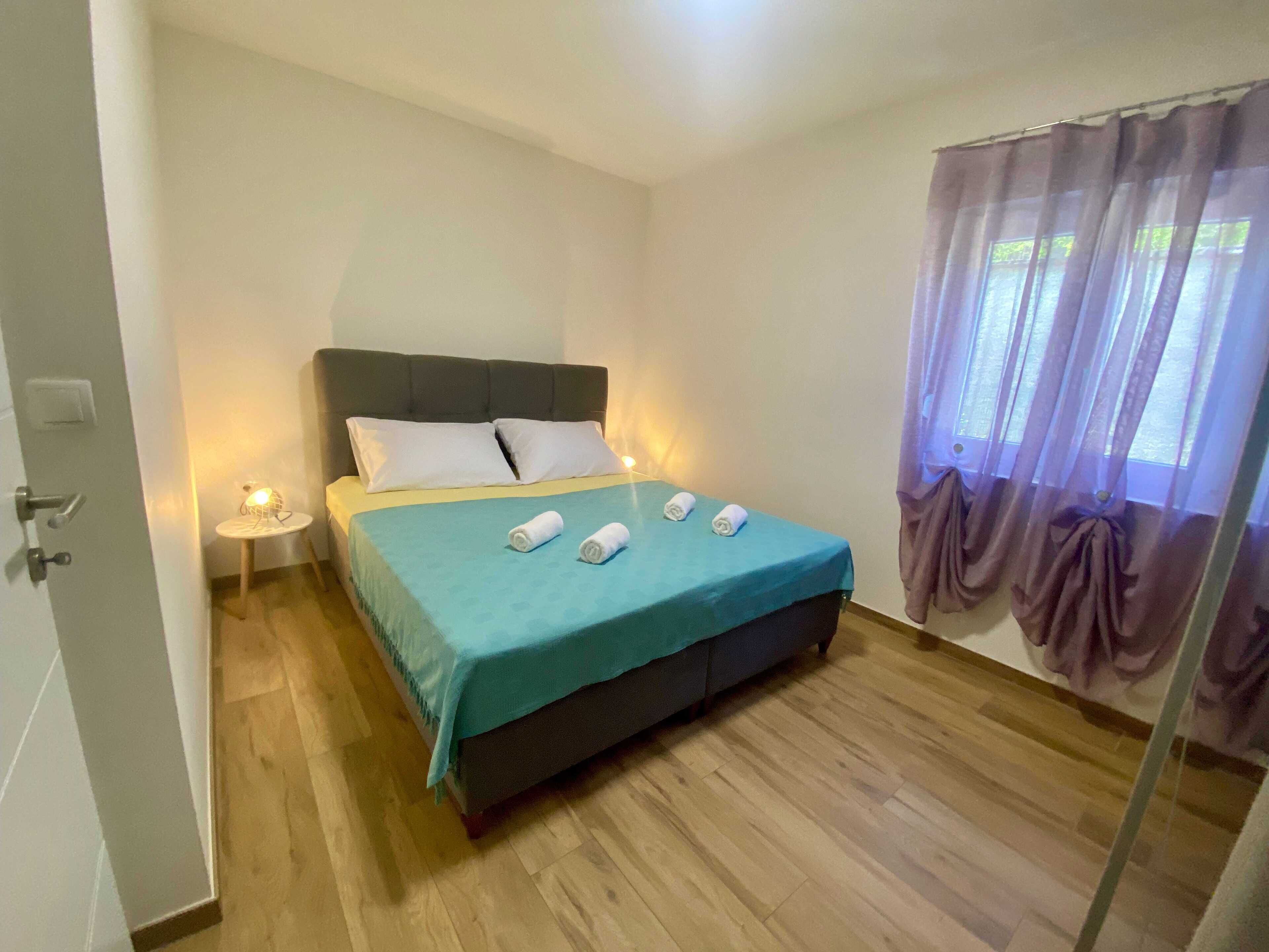 Een net opgemaakt bed met een blauwgroene sprei en twee opgerolde handdoeken, een nachtkastje met een lamp, en een raam met paarse gordijnen.