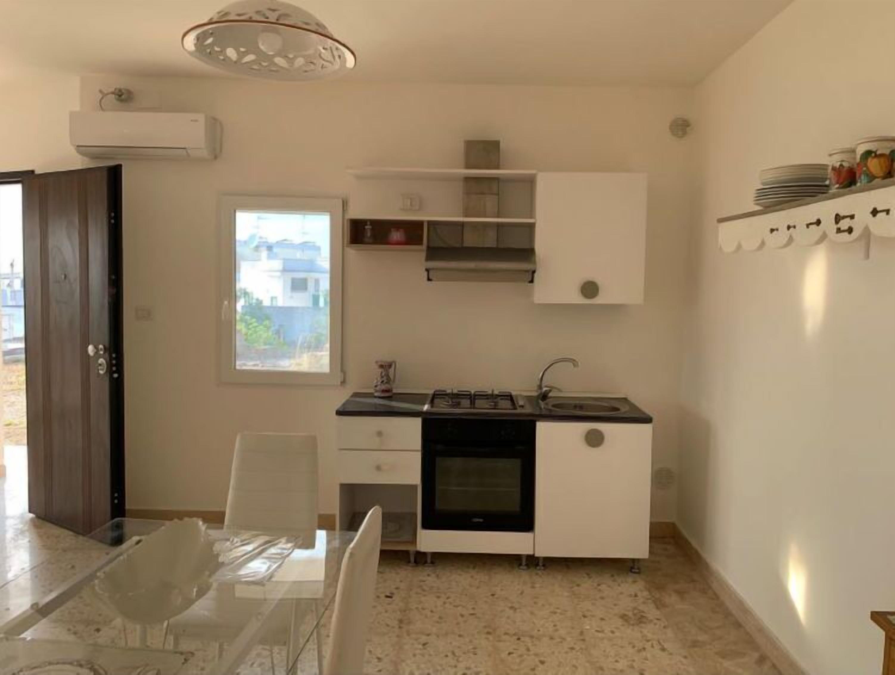 A compact kitchen with a gas stove, oven, and a small sink. There is a dining table with chairs, a window with a view of buildings, and a ceiling light fixture.