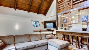 Una sala de estar moderna con un sofá modular, una zona de cocina con taburetes de bar y un techo de madera.