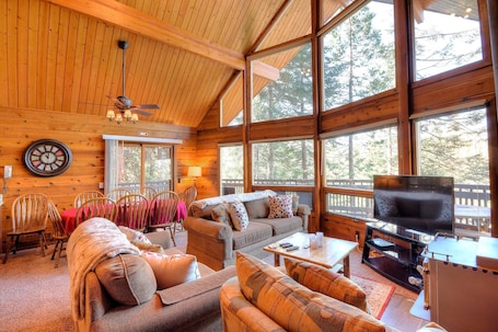 A cozy cabin interior with wooden walls, large windows, a ceiling fan, a TV, and a dining area with chairs and a table.