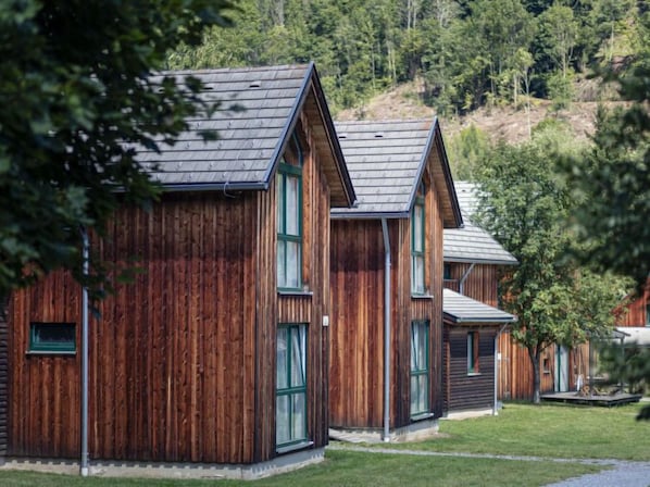 A row of wooden cabins with pitched roofs, set against a backdrop of trees and a hill.