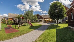 Un parque infantil con tobogán y columpios, una zona de estar cubierta y un edificio con mesas y sombrillas al aire libre.
