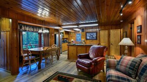 A cozy cabin interior with wooden walls, a dining area with chairs, a leather armchair, a plaid sofa, and a kitchen area with a refrigerator and sink. - Bear Foot Pines (CA 95389)