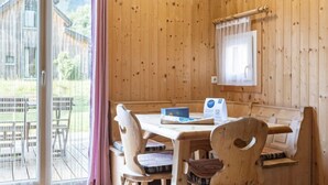 Eine Holzhütte mit Esstisch und Stühlen, einem mit Vorhängen drapierten Fenster und Blick auf ein Holzgebäude im Freien.