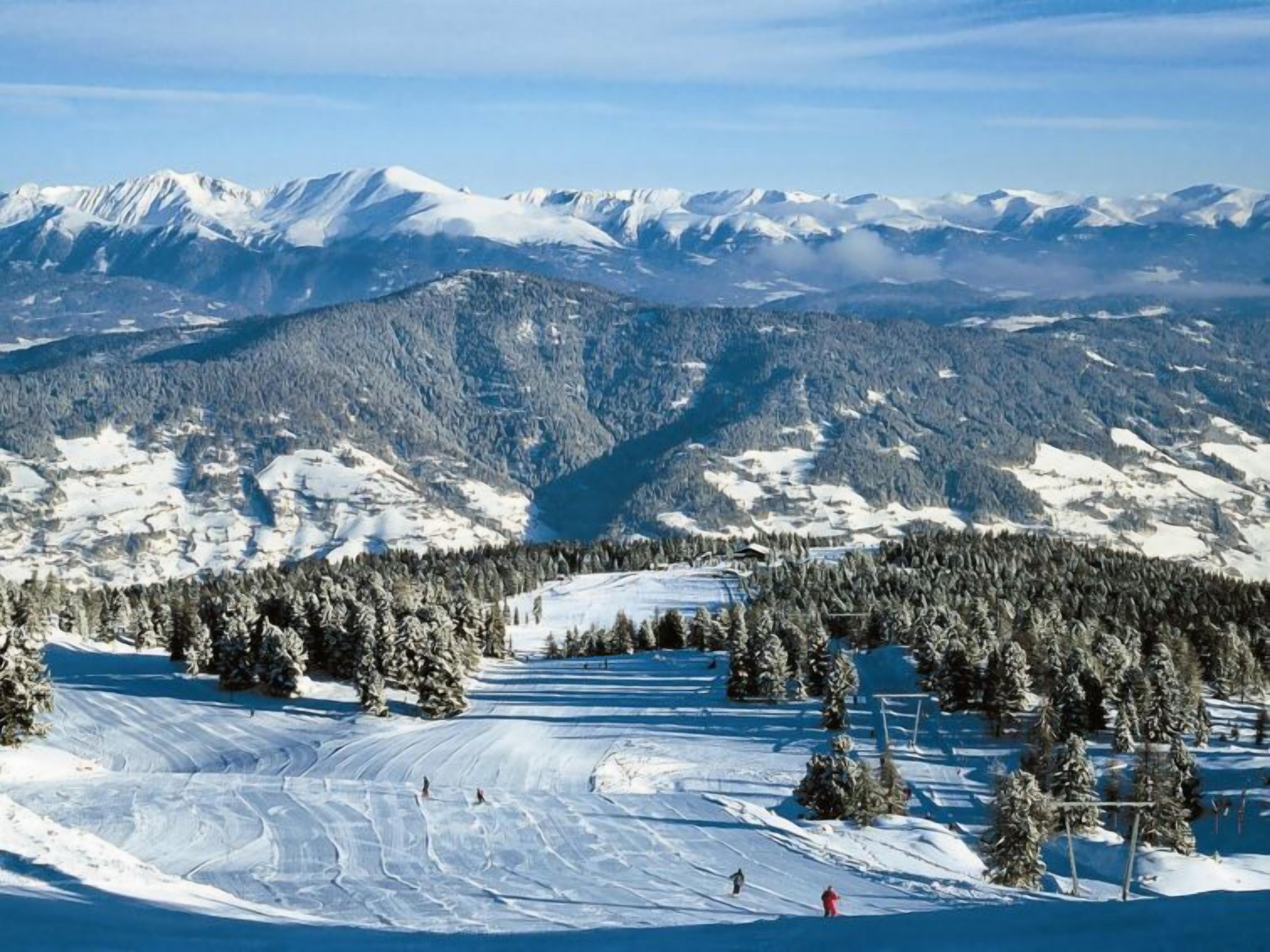 Une station de ski avec des pistes enneigées, une zone boisée et des montagnes qui se dessinent à l’horizon.