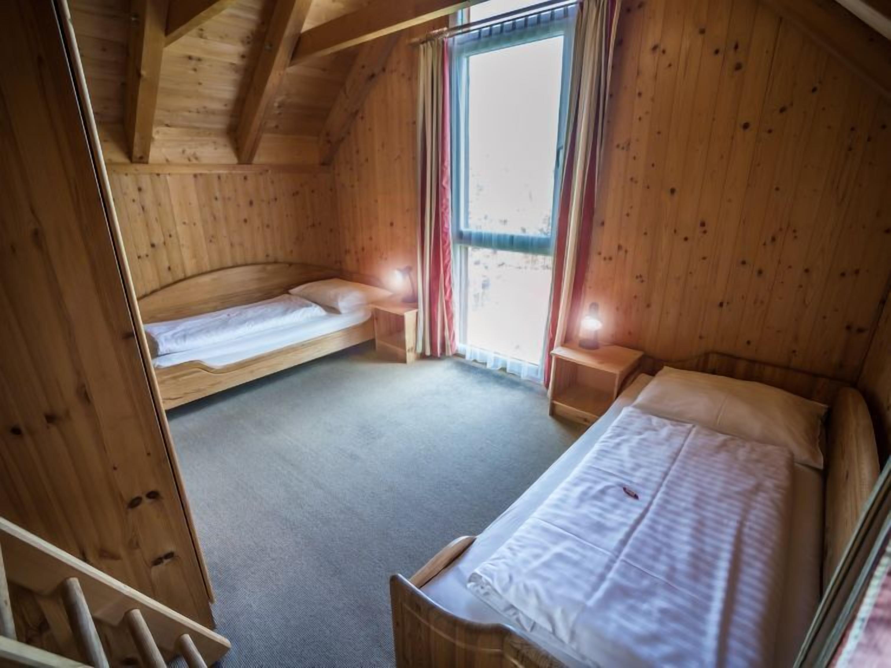 Une chambre dans un chalet en bois, avec deux lits simples, une petite table et une fenêtre avec des rideaux.