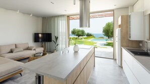 A modern kitchen with a large island, a dining area with a sofa, a TV, and a view of a pool and garden. - Luria Homes (Sithonia)
