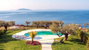 A swimming pool with a curved edge, surrounded by a grassy area and trees, with a view of the sea and distant mountains. - Luria Homes (Sithonia)