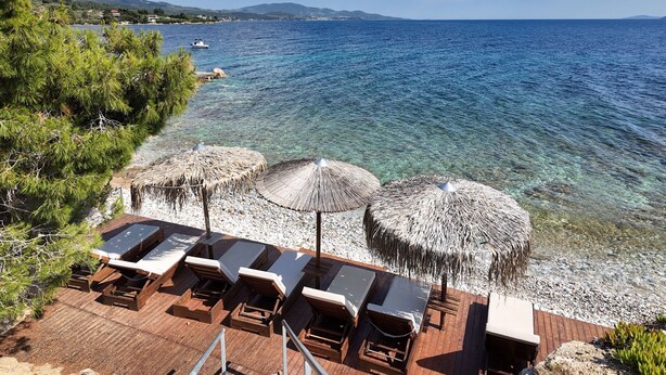 A wooden deck with lounge chairs and thatched umbrellas overlooking a clear blue sea with boats in the distance. - Luria Homes (Sithonia)