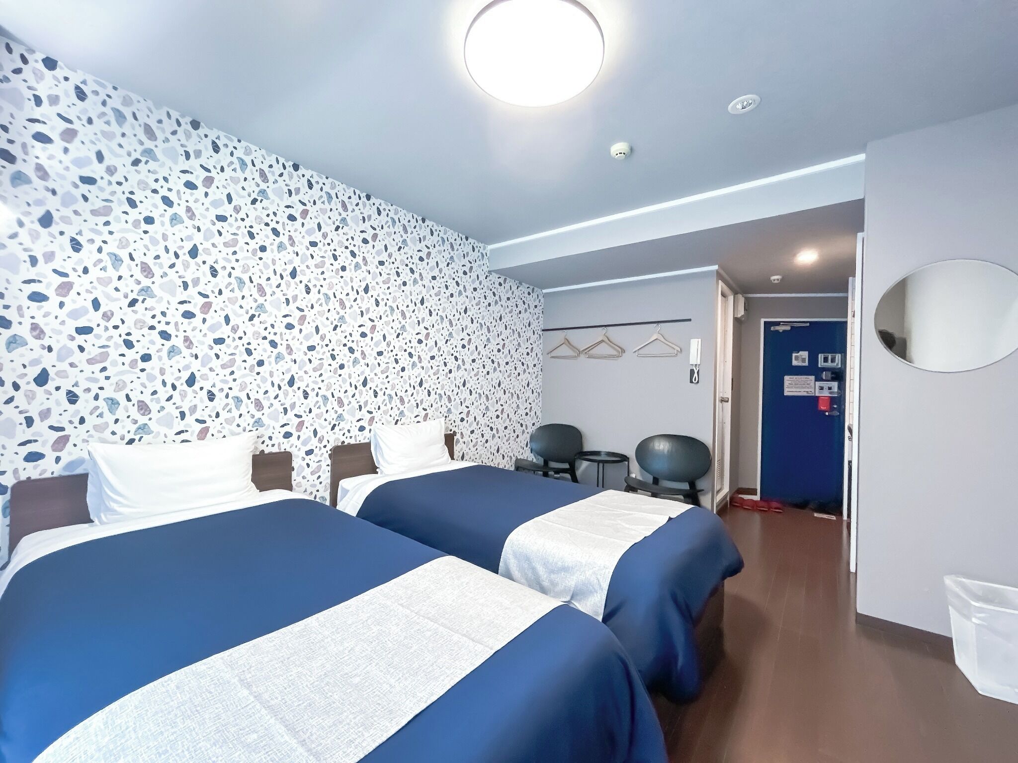 A hotel room with two beds, a wall with a terrazzo pattern, a round ceiling light, a door with a blue frame, and a small table with chairs.
