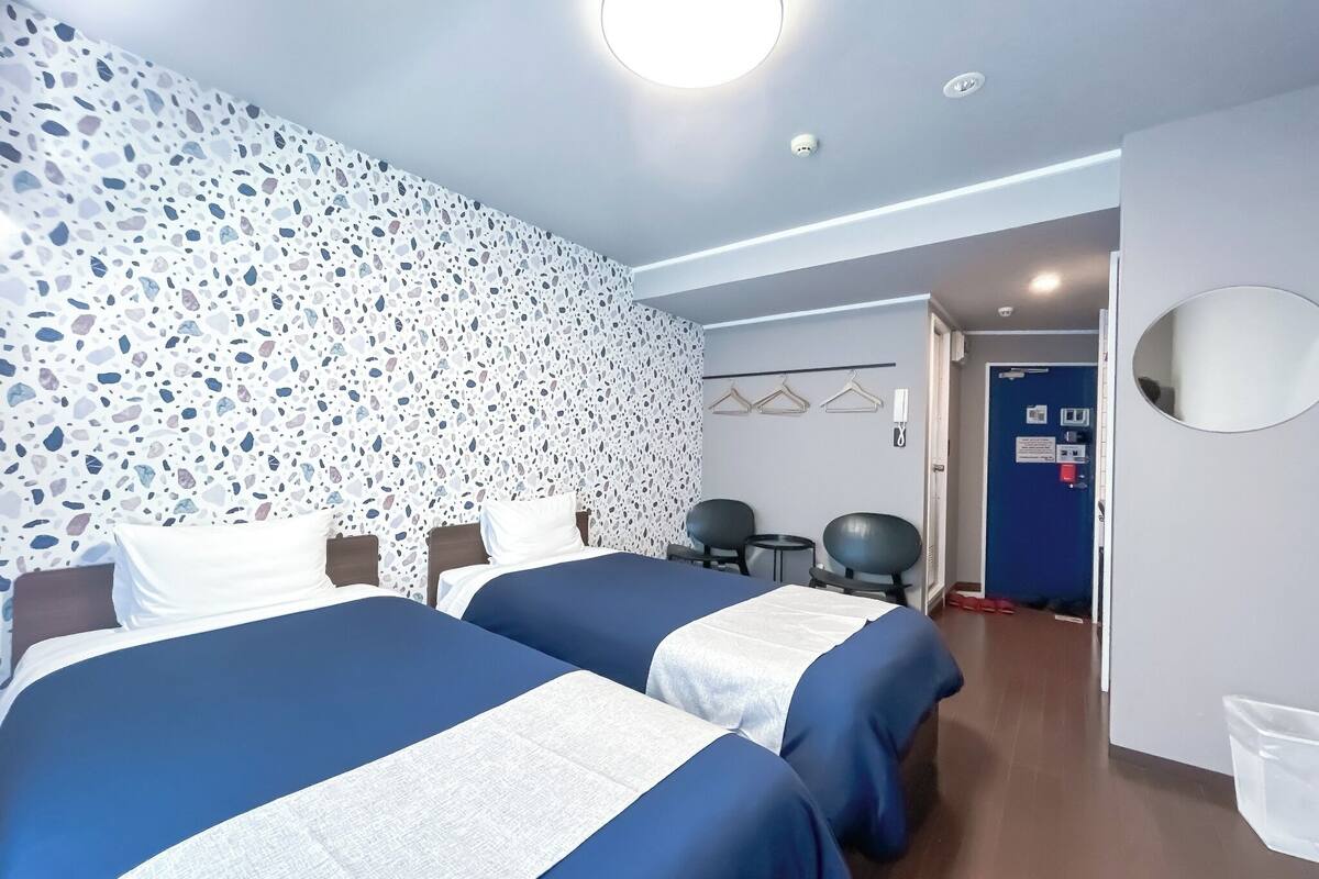 A hotel room with two beds, a wall with a terrazzo pattern, a round ceiling light, a door with a blue frame, and a small table with chairs.
