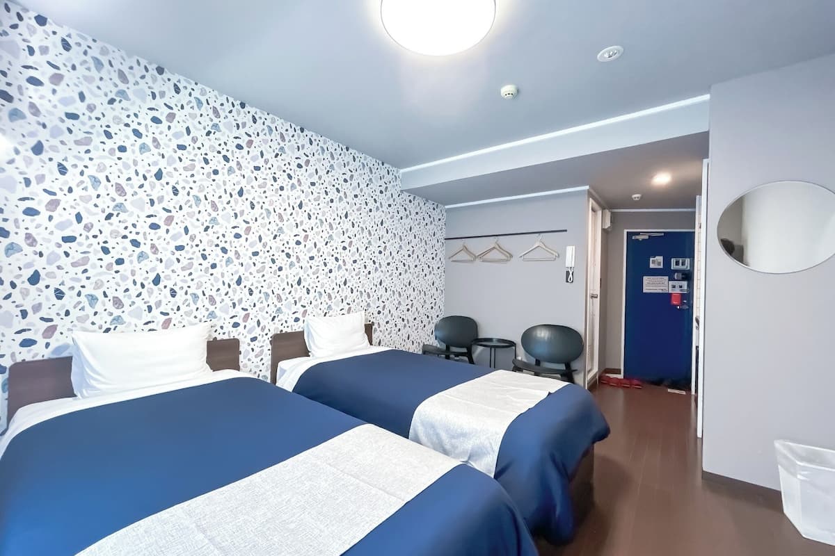 A hotel room with two beds, a wall with a terrazzo pattern, a round ceiling light, a door with a blue frame, and a small table with chairs.